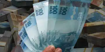 Pessoa segurando notas de cem reais em frente a dinheiro