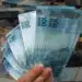 Pessoa segurando notas de cem reais em frente a dinheiro