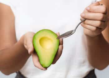 Foto em plano médio e em close, focada nas mãos de uma pessoa com pele de tom médio, usando uma camiseta branca. A mão esquerda segura firmemente uma metade de abacate maduro, verde brilhante, com a cavidade central do caroço vazia. A mão direita aproxima uma colher de sobremesa de metal prateado da polpa da fruta, como se fosse retirar uma porção. O fundo é neutro e desfocado.