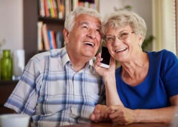 Casal de idosos sorrindo alegremente enquanto compartilham uma ligação no celular em um ambiente doméstico.