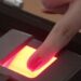 Dedo com esmalte vermelho pressionado em leitor biométrico com luz vermelha