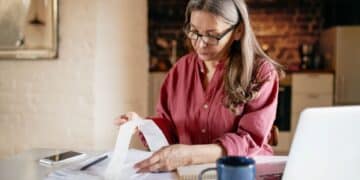 Uma mulher de óculos e blusa rosa sentada à mesa, analisando atentamente uma longa nota fiscal de papel. À sua frente, há documentos, um notebook aberto, um smartphone e uma caneca azul. O ambiente sugere um escritório doméstico aconchegante.