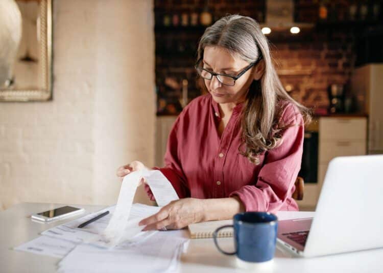 Uma mulher de óculos e blusa rosa sentada à mesa, analisando atentamente uma longa nota fiscal de papel. À sua frente, há documentos, um notebook aberto, um smartphone e uma caneca azul. O ambiente sugere um escritório doméstico aconchegante.