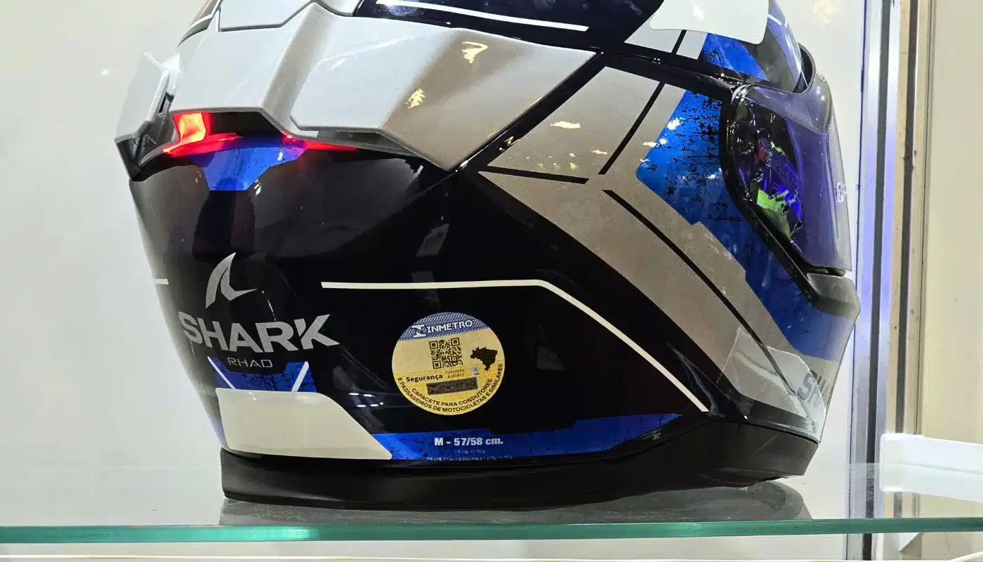 Close da parte traseira de um capacete de luxo da marca Shark, nas cores azul, prata e preto. No centro da base, destaca-se o novo selo digital do Inmetro, com um QR Code centralizado e o mapa do Brasil à direita, aplicado sobre a superfície brilhante do equipamento.