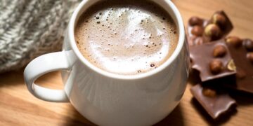 Caneca branca arredondada com chocolate quente cremoso e espuma no topo, servida sobre uma tábua de madeira redonda. Ao lado, pedaços de chocolate com avelãs e uma manta de lã cinza ao fundo.