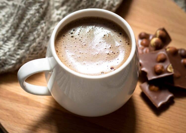 Caneca branca arredondada com chocolate quente cremoso e espuma no topo, servida sobre uma tábua de madeira redonda. Ao lado, pedaços de chocolate com avelãs e uma manta de lã cinza ao fundo.