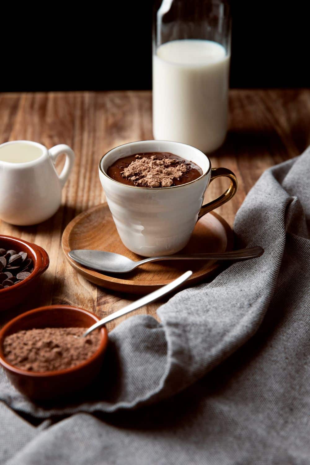Xícara branca com detalhes dourados cheia de chocolate quente cremoso e polvilhada com cacau em pó. Ao redor, sobre uma mesa de madeira, há uma garrafa de leite, pequenas tigelas com gotas de chocolate e cacau, uma colher de metal e um pano cinza.