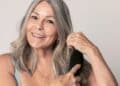 Retrato de uma mulher madura e sorridente, com cabelos grisalhos longos e ondulados, escovando os fios com uma escova preta oval. Ela possui pele clara com algumas sardas, usa brincos dourados e anéis delicados. O fundo é neutro e claro, destacando a vitalidade e o brilho natural dos cabelos prateados.