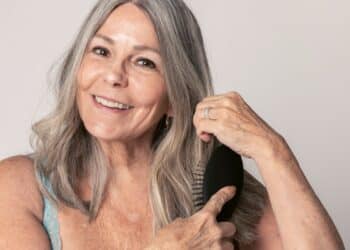 Retrato de uma mulher madura e sorridente, com cabelos grisalhos longos e ondulados, escovando os fios com uma escova preta oval. Ela possui pele clara com algumas sardas, usa brincos dourados e anéis delicados. O fundo é neutro e claro, destacando a vitalidade e o brilho natural dos cabelos prateados.
