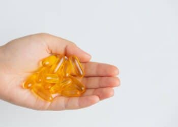 Palma de uma mão aberta segurando cerca de quinze cápsulas gelatinosas e transparentes de cor amarela intensa, representando suplementos de vitamina D. Fundo branco liso e iluminação clara.