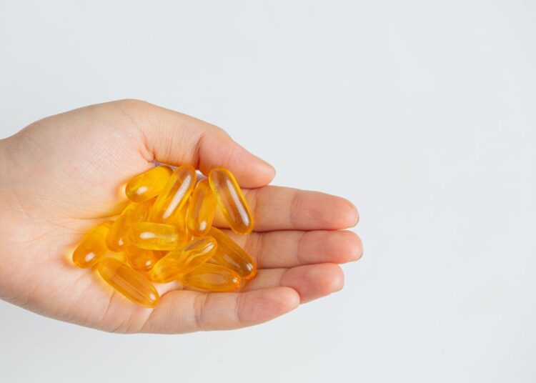 Palma de uma mão aberta segurando cerca de quinze cápsulas gelatinosas e transparentes de cor amarela intensa, representando suplementos de vitamina D. Fundo branco liso e iluminação clara.
