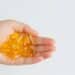 Palma de uma mão aberta segurando cerca de quinze cápsulas gelatinosas e transparentes de cor amarela intensa, representando suplementos de vitamina D. Fundo branco liso e iluminação clara.