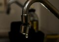 Close em uma torneira metálica de cozinha com uma gota de água prestes a cair. O fundo está escurecido e desfocado, destacando o desperdício ou a economia de água.