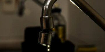 Close em uma torneira metálica de cozinha com uma gota de água prestes a cair. O fundo está escurecido e desfocado, destacando o desperdício ou a economia de água.