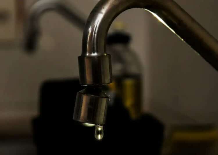 Close em uma torneira metálica de cozinha com uma gota de água prestes a cair. O fundo está escurecido e desfocado, destacando o desperdício ou a economia de água.