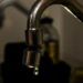 Close em uma torneira metálica de cozinha com uma gota de água prestes a cair. O fundo está escurecido e desfocado, destacando o desperdício ou a economia de água.