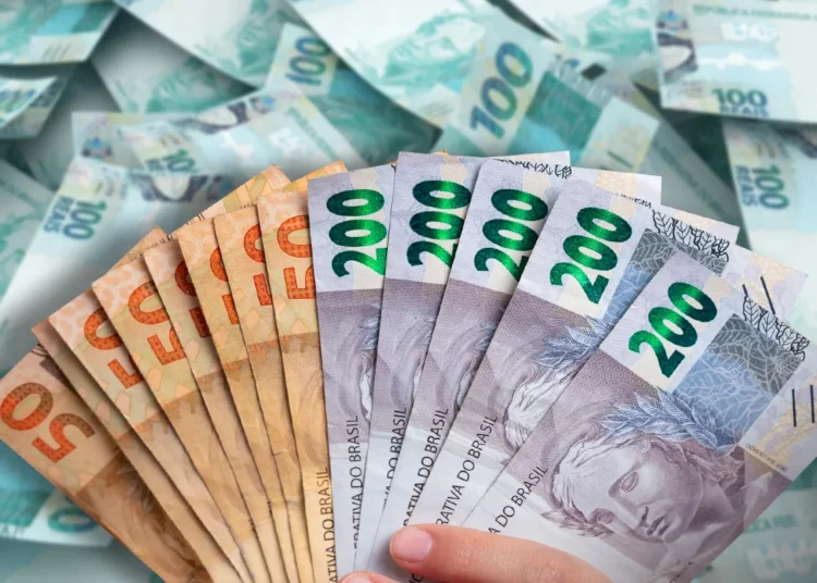 Mão segurando leque de notas de R$ 200 e R$ 50 com várias cédulas de R$ 100 ao fundo