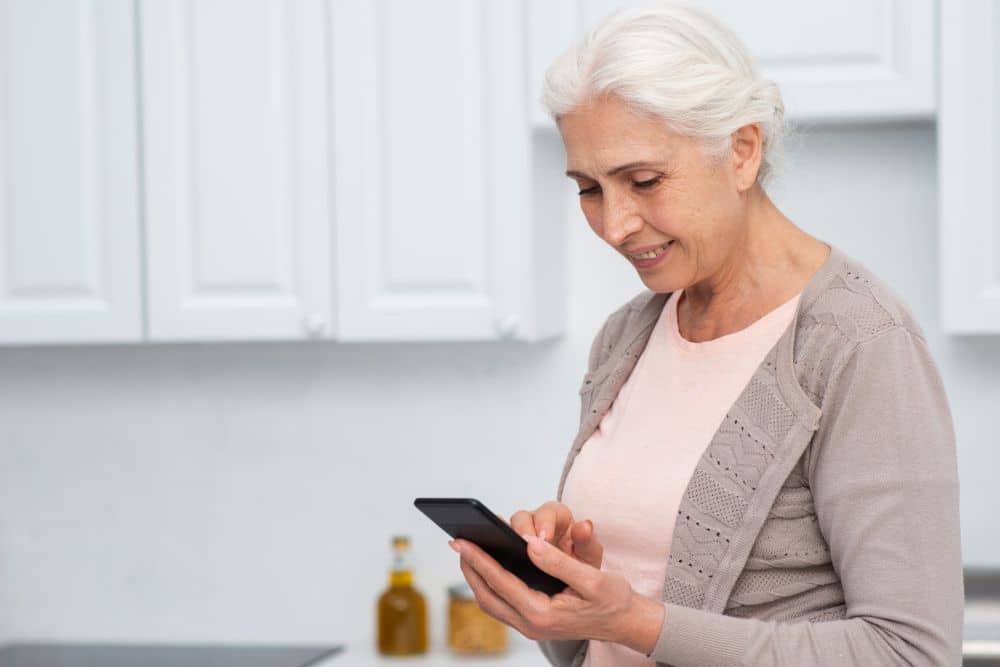 Mulher idosa de cabelos brancos, vestindo um cardigã bege, sorrindo enquanto digita uma mensagem no smartphone em uma cozinha clara.