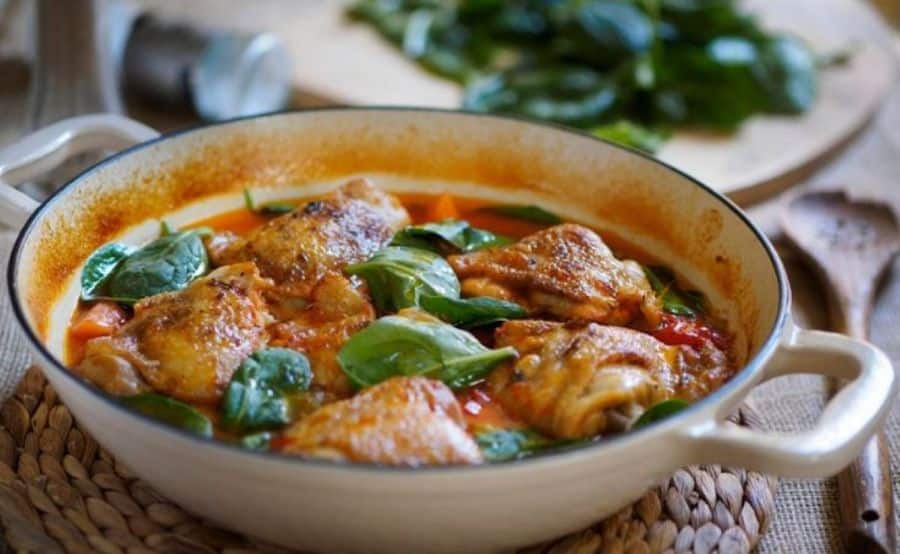 Uma panela de cerâmica clara com pedaços de frango dourados ao molho, decorados com folhas frescas de ora-pro-nóbis verde-escuras. Ao fundo, uma tábua com mais folhas da planta e uma colher de pau, sobre um descanso de palha.