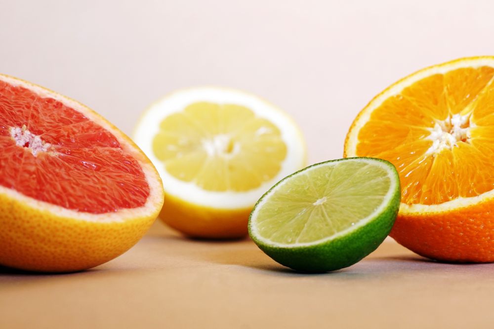 Close-up de quatro metades de frutas cítricas diferentes, exibindo suas polpas suculentas: uma toranja (rosa), uma laranja (alaranjada), um limão siciliano (amarelo) e um limão taiti (verde) em primeiro plano. O fundo é neutro e de cor clara.