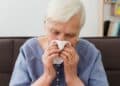 Mulher idosa de cabelos brancos, vestindo uma blusa azul, com expressão de abatimento e olhos fechados enquanto usa um lenço de papel branco para cobrir o nariz e a boca. Ela parece estar resfriada ou com gripe, ilustrando os sintomas de infecções respiratórias.