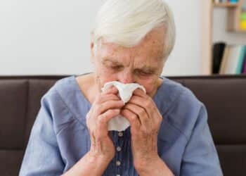 Mulher idosa de cabelos brancos, vestindo uma blusa azul, com expressão de abatimento e olhos fechados enquanto usa um lenço de papel branco para cobrir o nariz e a boca. Ela parece estar resfriada ou com gripe, ilustrando os sintomas de infecções respiratórias.
