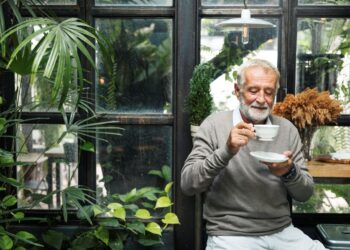 Homem idoso de barba grisalha e suéter cinza sorrindo enquanto segura uma xícara de café branca em um ambiente iluminado com plantas ao fundo.
