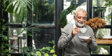 Homem idoso de barba grisalha e suéter cinza sorrindo enquanto segura uma xícara de café branca em um ambiente iluminado com plantas ao fundo.