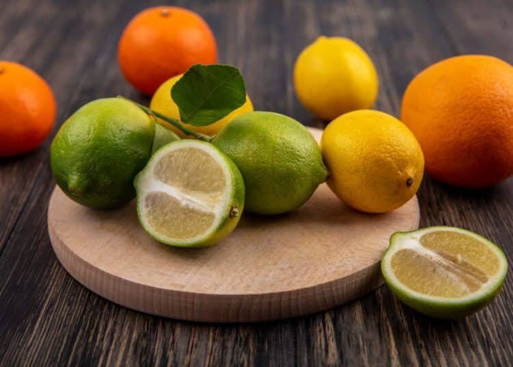 Fotografia de estúdio colorida sobre uma mesa de madeira escura. Centralizada sobre uma tábua de cortar redonda de madeira clara, estão três limões Taiti (casca verde), um deles cortado ao meio revelando a polpa, e um limão siciliano (casca amarela). Ao fundo, espalhados diretamente sobre a mesa, estão uma laranja inteira (casca laranja), outra mexerica e mais dois limões amarelos. Um pedaço pequeno de limão verde cortado está na mesa, em primeiro plano. A iluminação é suave e natural.