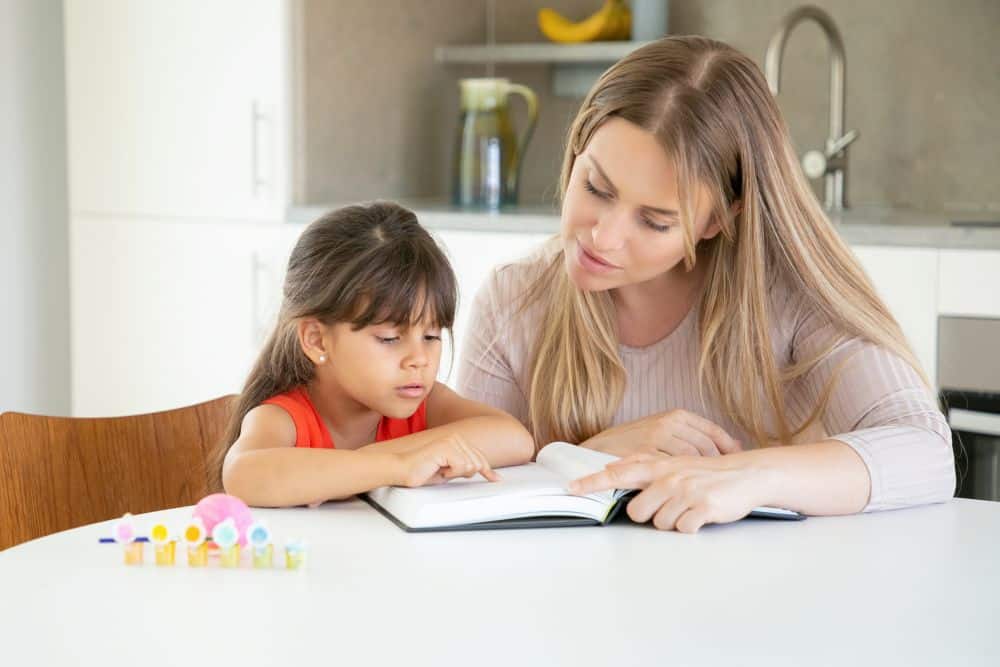 Uma mulher loira sentada à mesa de uma cozinha, orientando uma menina pequena na leitura de um livro. A mãe aponta para as páginas enquanto a criança acompanha atentamente. O ambiente é organizado e iluminado.