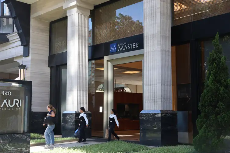 Fotografia da fachada externa de uma agência do Banco Master, localizada no Edifício Auri, na Avenida Faria Lima. A imagem mostra a entrada principal com portas de vidro, colunas brancas de estilo clássico e o letreiro azul e branco com a logomarca do banco. Funcionários uniformizados e pedestres circulam em frente ao prédio em um dia ensolarado.