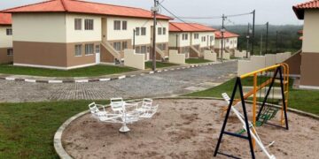 Vista de um conjunto habitacional do programa Minha Casa, Minha Vida com casas geminadas de dois pavimentos, cores bege e marrom, telhado de barro e um parquinho infantil em primeiro plano.