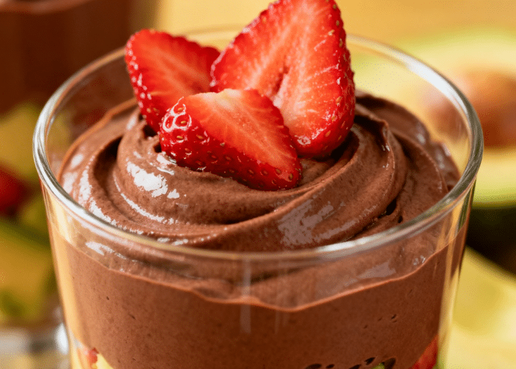 Um copo de vidro transparente com uma sobremesa em camadas: na base, cubos de abacate e morangos frescos; acima, um mousse de chocolate aerado; no topo, fatias de morango fresco. Ao fundo, um segundo copo e metade de um abacate, sobre uma superfície dourada e desfocada.