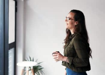 Mulher jovem sorrindo e olhando pela janela enquanto segura um café, ilustrando o hábito saudável de falar sozinho e organizar pensamentos segundo a psicologia.