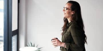 Mulher jovem sorrindo e olhando pela janela enquanto segura um café, ilustrando o hábito saudável de falar sozinho e organizar pensamentos segundo a psicologia.