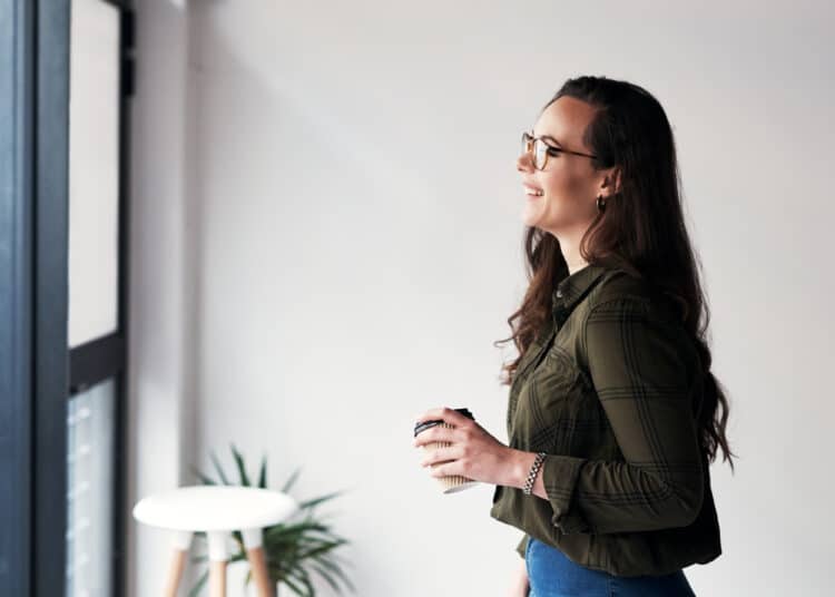 Mulher jovem sorrindo e olhando pela janela enquanto segura um café, ilustrando o hábito saudável de falar sozinho e organizar pensamentos segundo a psicologia.