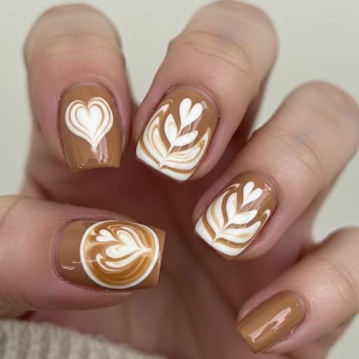 Close das unhas de uma mão com formato quadrado e médio. A esmaltação é em tom marrom caramelo cremoso, decorada com desenhos brancos que imitam a "latte art" de cafeterias: corações e folhas estilizadas que lembram a espuma do cappuccino.