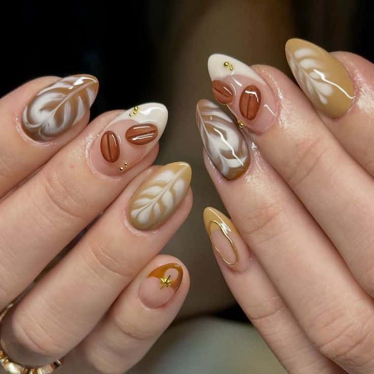 Duas mãos com unhas amendoadas exibindo uma nail art de luxo. A decoração inclui grãos de café em relevo 3D na cor marrom, desenhos de latte art em branco e tons caramelo, e pequenos detalhes em ouro metálico. A base é nude translúcida com acabamento em gel brilhante.