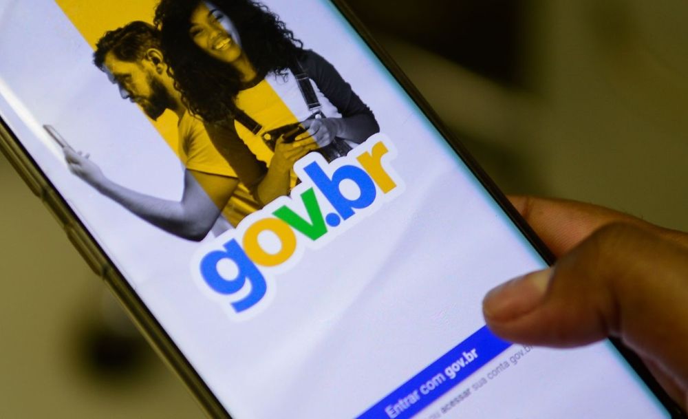Mão segurando um smartphone exibindo a tela inicial de login do aplicativo Gov.br, com o logotipo colorido em destaque sobre fundo branco e azul.