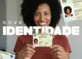 Close de uma mulher negra sorridente, com cabelo cacheado volumoso e vestindo uma blusa coral, segurando em primeiro plano a Nova Carteira de Identidade Nacional (CIN) em papel. O texto "NOVA IDENTIDADE" está sobreposto em destaque em letras brancas sobre ela. Outros exemplares do documento aparecem desfocados nos cantos superiores e inferiores, ilustrando o novo modelo.