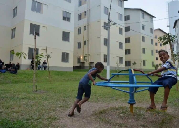 Crianças brincando em um gira-gira no pátio de um conjunto habitacional do programa Minha Casa, Minha Vida, com prédios novos e modernos ao fundo.