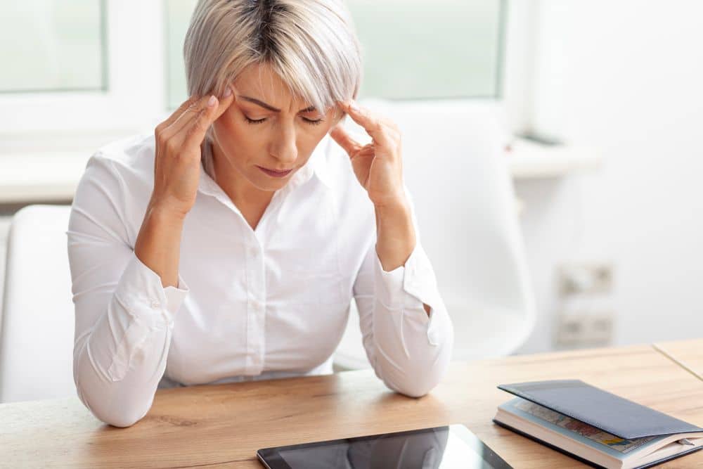 Mulher de cabelos curtos platinados sentada à mesa de trabalho, com os olhos fechados e as mãos nas têmporas, demonstrando cansaço ou lapso de memória.