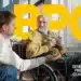 Idoso em cadeira de rodas segurando um copo de água e conversando com um cuidador, com a sigla BPC e o texto Benefício de Prestação Continuada em destaque amarelo ao fundo.