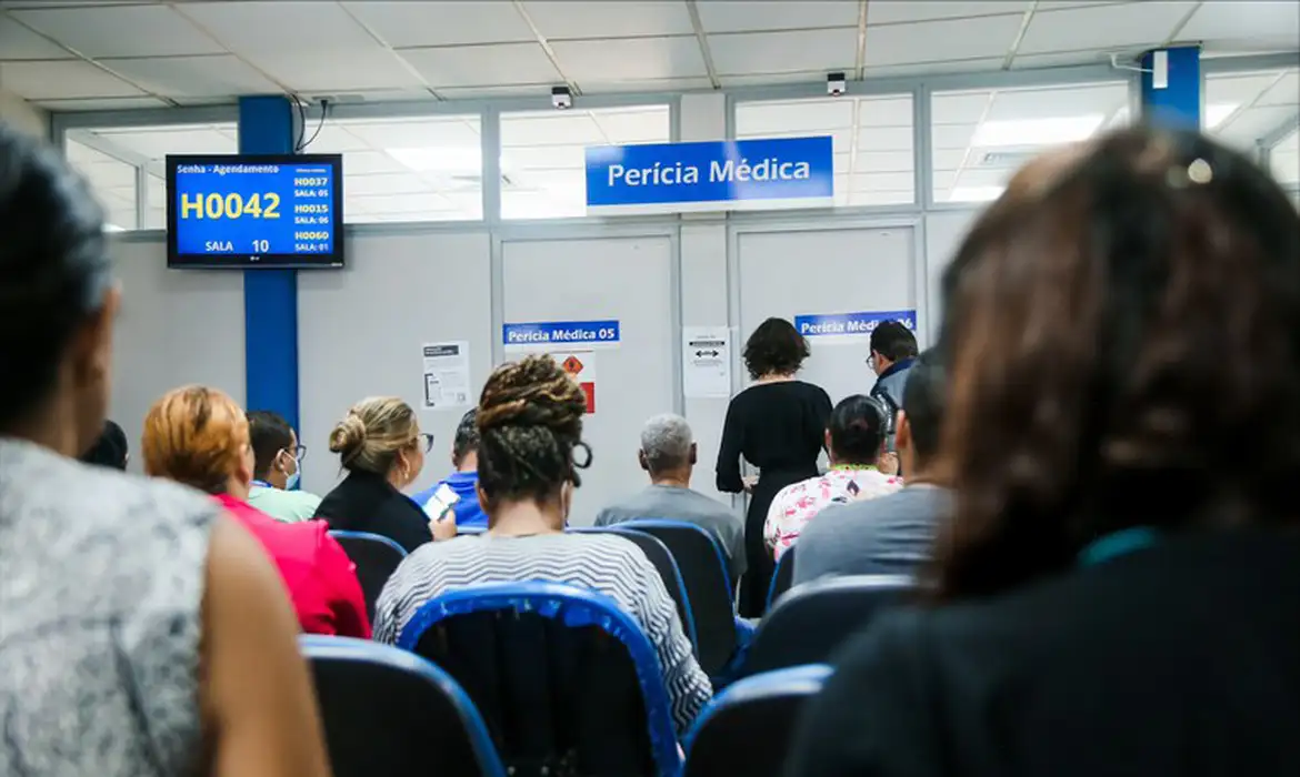 Vista interna de uma sala de espera do INSS com pessoas sentadas de costas aguardando atendimento. Ao fundo, portas com placas indicando "Perícia Médica" e um painel eletrônico azul exibindo a senha H0042.