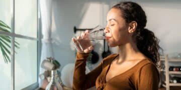 Mulher jovem bebendo um copo d’água em uma cozinha iluminada pela manhã, ilustrando os benefícios de se hidratar em jejum ao acordar para a saúde digestiva e energia.