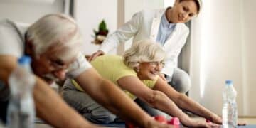 Uma instrutora de jaleco branco sorri enquanto orienta uma mulher idosa de cabelos loiros e camiseta amarela em um exercício de alongamento no chão. Ao lado, um homem idoso também pratica o movimento em um tapete de yoga. Garrafas de água e halteres rosas estão próximos no chão.