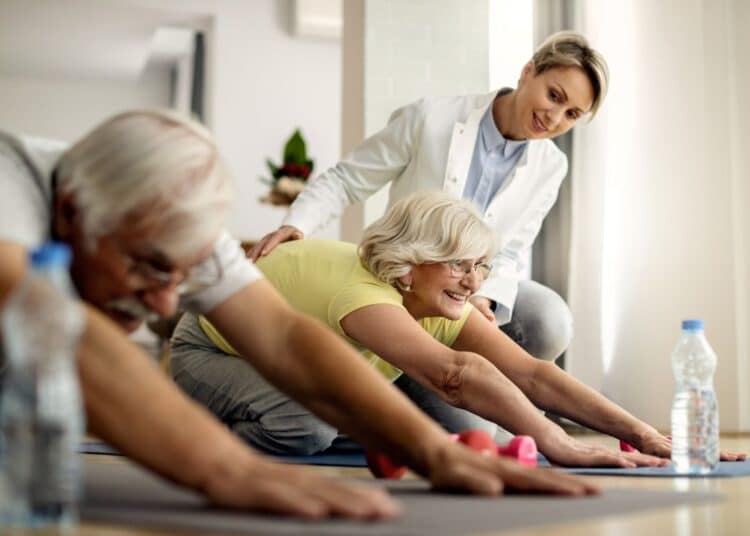 Uma instrutora de jaleco branco sorri enquanto orienta uma mulher idosa de cabelos loiros e camiseta amarela em um exercício de alongamento no chão. Ao lado, um homem idoso também pratica o movimento em um tapete de yoga. Garrafas de água e halteres rosas estão próximos no chão.
