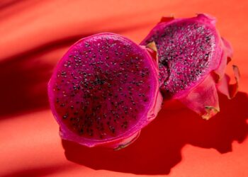 Close detalhado de uma pitaia de polpa rosa vibrante cortada ao meio, exibindo suas pequenas sementes pretas sobre um fundo de cor sólida. A imagem destaca a textura suculenta da fruta, associada a benefícios para a saúde das artérias.