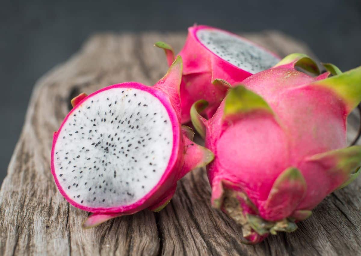 Três pitaias de casca rosa vibrante sobre uma superfície de madeira rústica. Uma delas está cortada ao meio, revelando a polpa branca densamente salpicada de sementes pretas. A imagem foca na variedade branca, rica em fibras e magnésio.