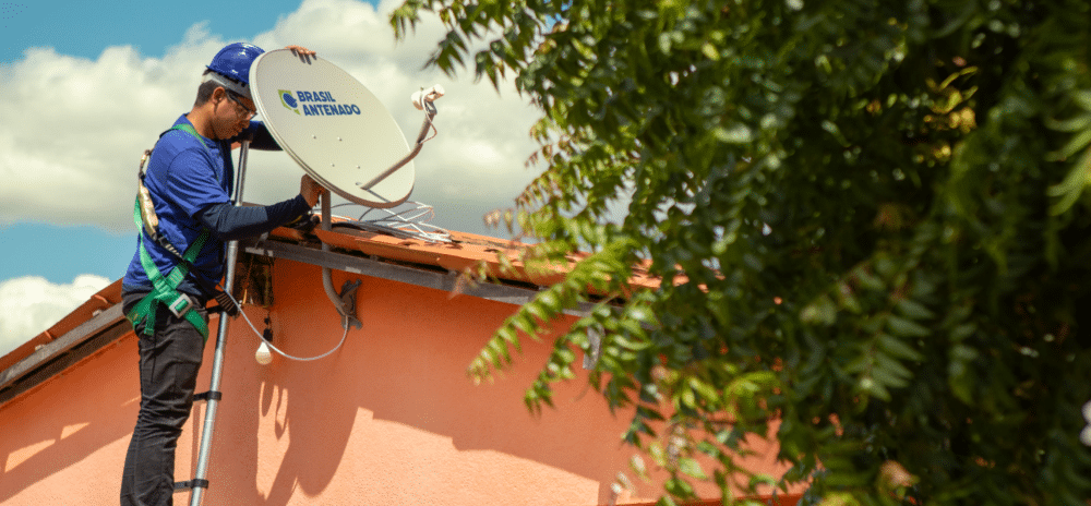 Um instalador de camisa azul e capacete azul sobre uma escada, segurando uma antena parabólica digital branca no telhado de uma casa. A antena tem o logotipo "BRASIL ANTENADO". Uma árvore com folhas verdes está em primeiro plano no canto direito. O céu está azul com algumas nuvens.